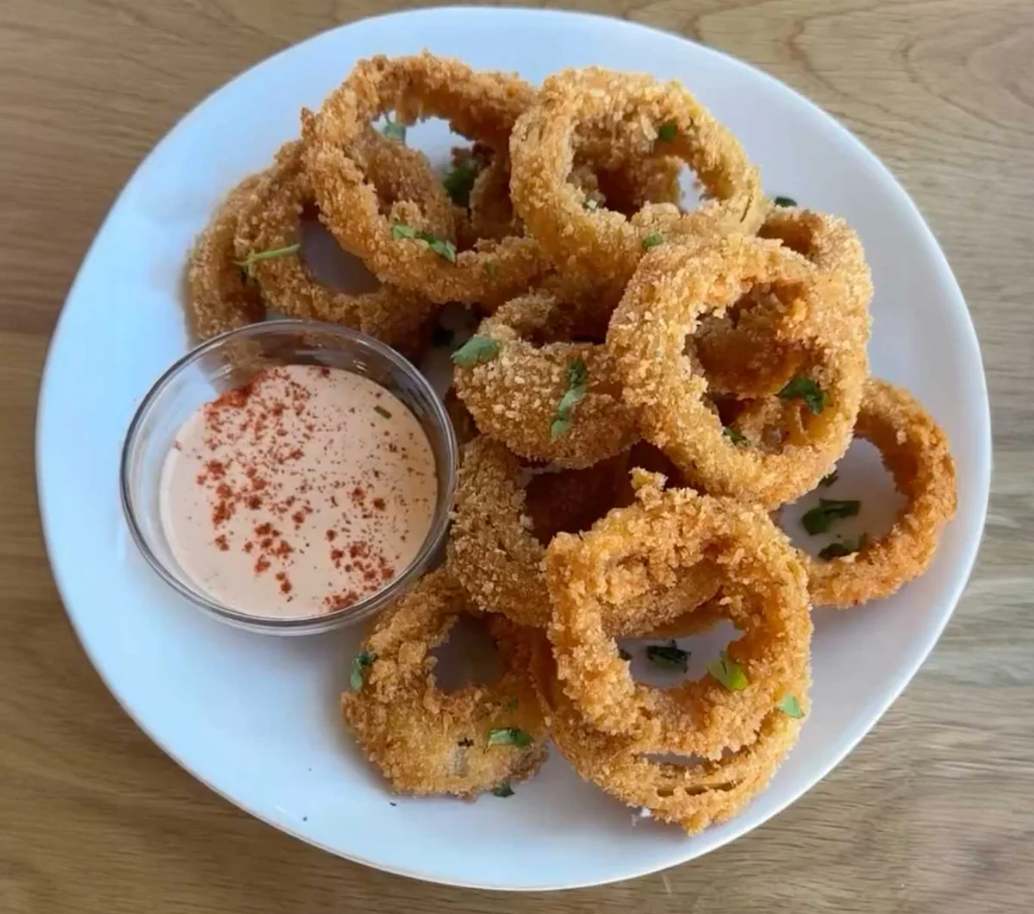 A pile of golden brown crispy homemade onion rings served with sriracha aioli, fried to perfection in a Pink Lynnee non-stick skillet.