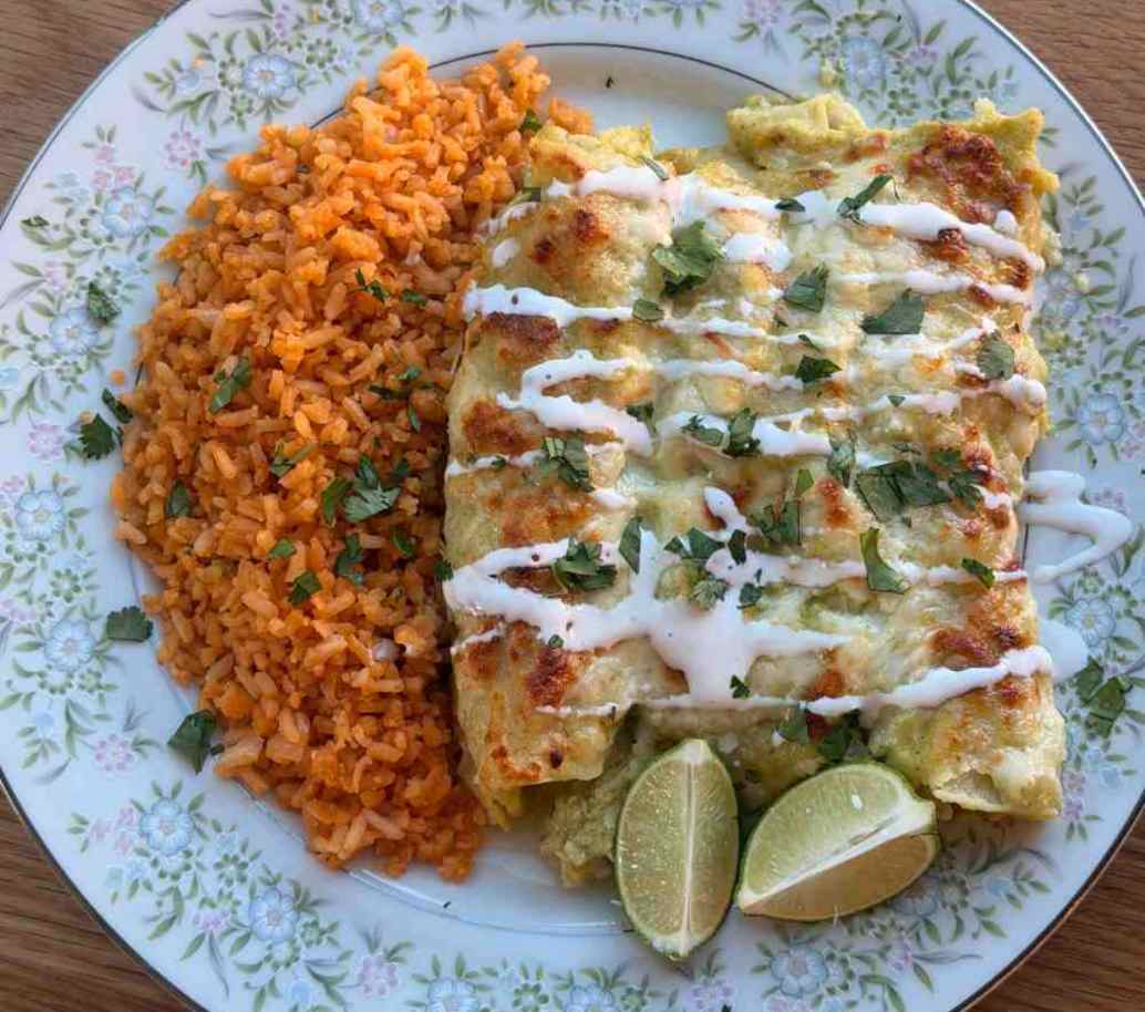 A close-up plate of creamy green chicken enchiladas topped with melted Monterey Jack cheese, queso fresco, and cilantro, served with rice.